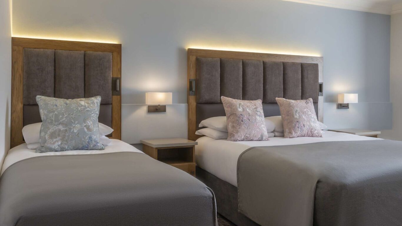 A hotel room with two beds, each with gray upholstered headboards, neutral bedding, floral pillows, and wall-mounted bedside lamps.