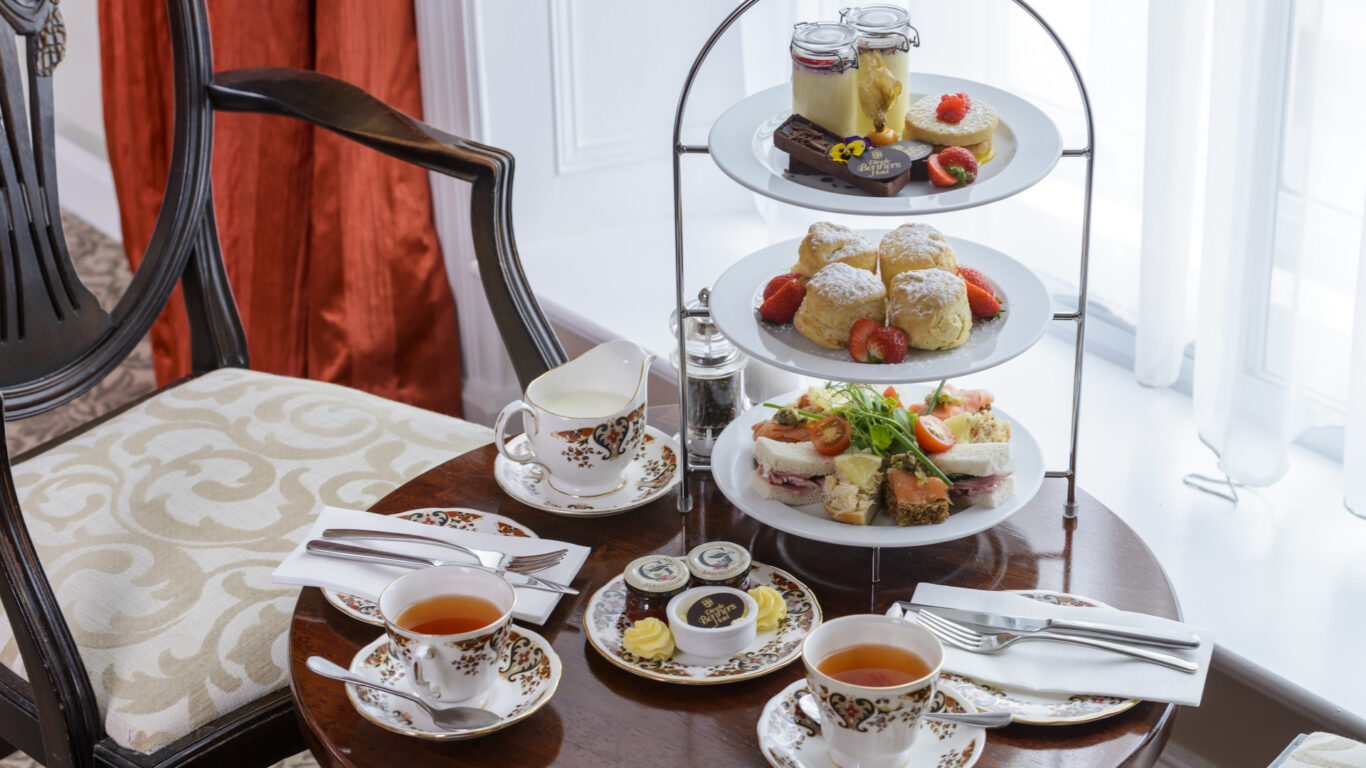 A round wooden table set for afternoon tea with two cups of tea, a tiered tray of pastries, scones, sandwiches, jam jars, and cream, next to upholstered chairs by a window.