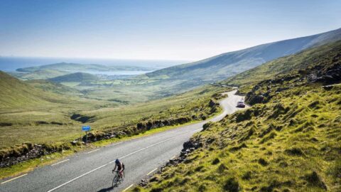 A cyclist rides uphill on a winding road through a green, hilly landscape with distant water and a clear blue sky.