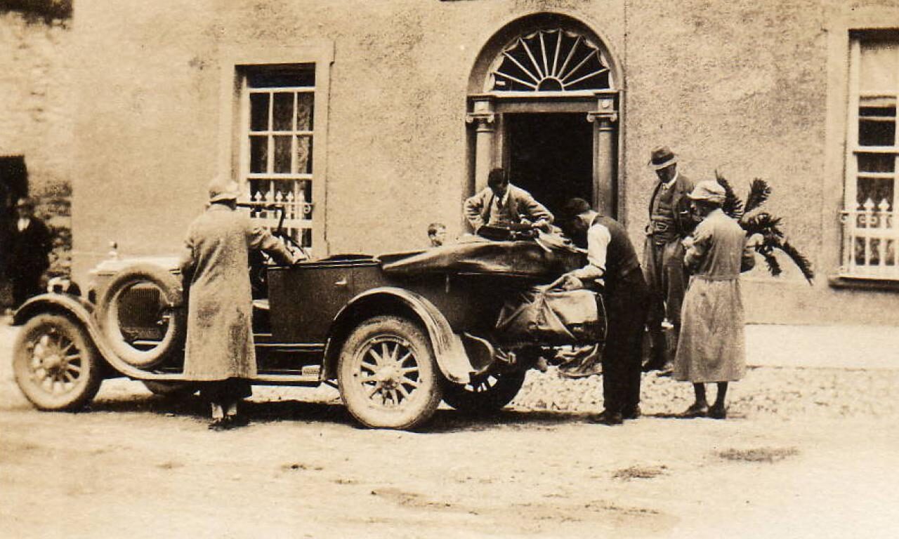 Several people stand around a vintage car parked outside a building labeled 
