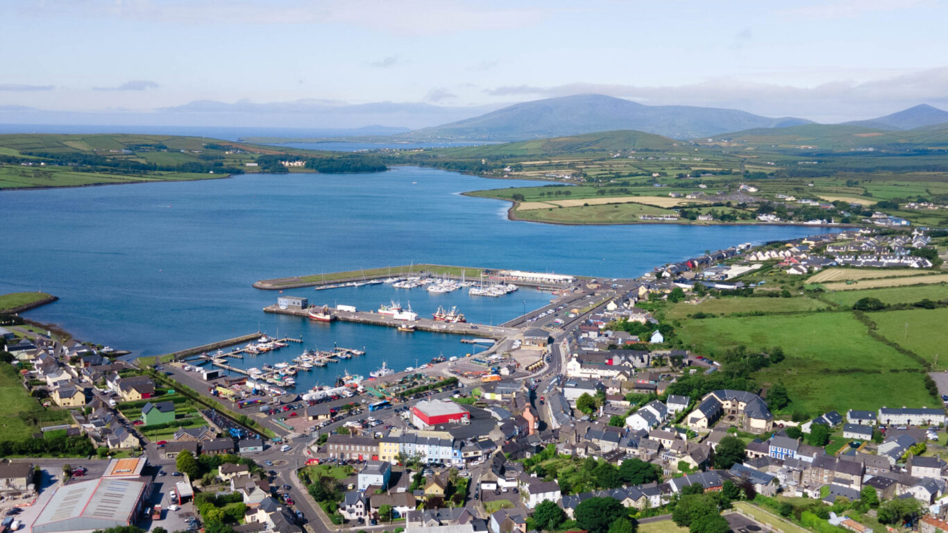 Aerial view of a coastal town with a marina, boats docked, green fields, and distant hills under a blue sky with scattered clouds.