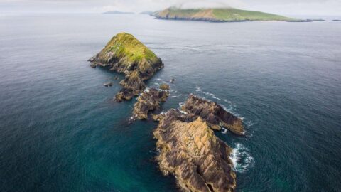 Aerial view of rocky, green-topped islands in the ocean with a larger, mist-covered island in the background under a cloudy sky.