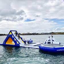 Large inflatable water obstacle course floating on the sea with several people climbing and playing on it under a cloudy sky.