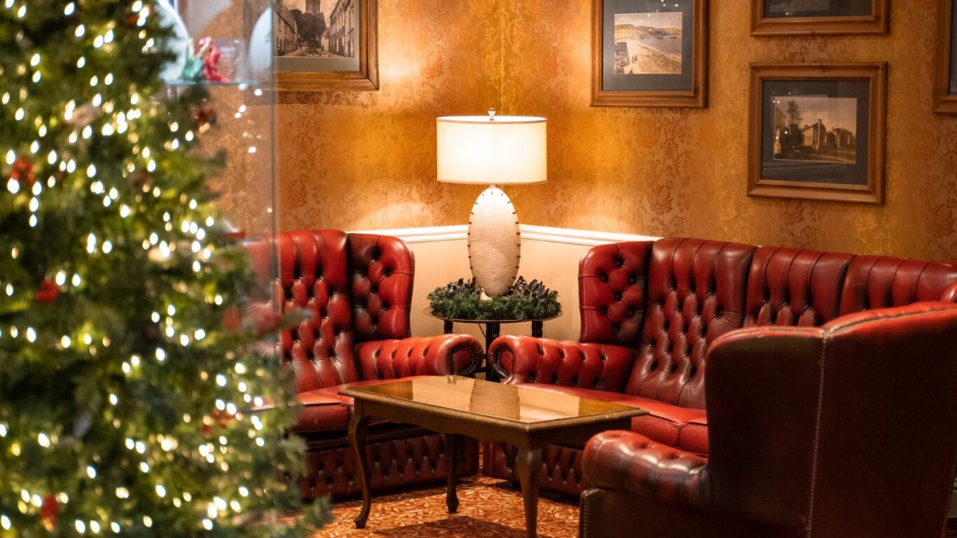 A warmly lit lounge features red leather furniture, a wooden coffee table, framed pictures on patterned walls, and a decorated Christmas tree in the foreground.