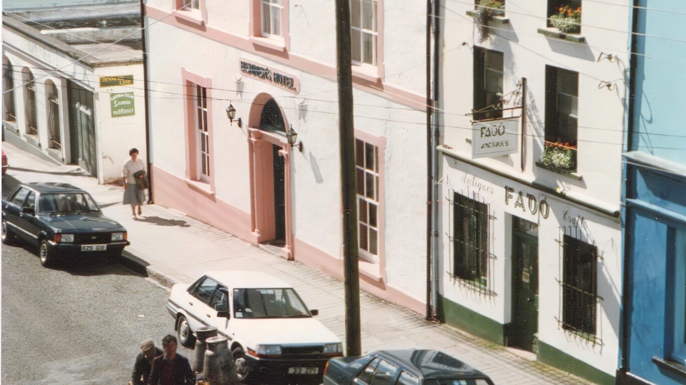 A horse-drawn cart travels down a street lined with parked cars and buildings, including a hotel and a pub, on a sunny day.