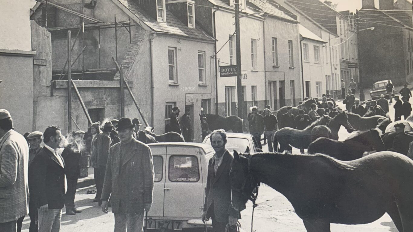 Black and white photo of a busy street scene with people, cars, and horses; buildings line both sides of the street and a crowd gathers in the background.