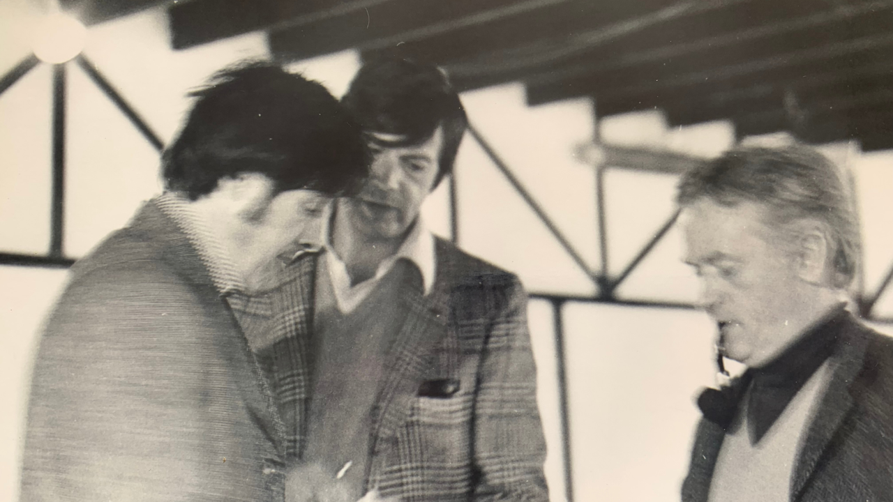 Three men in jackets and sweaters stand together indoors, engaged in conversation or an activity, with a blurred background of beams and geometric patterns.