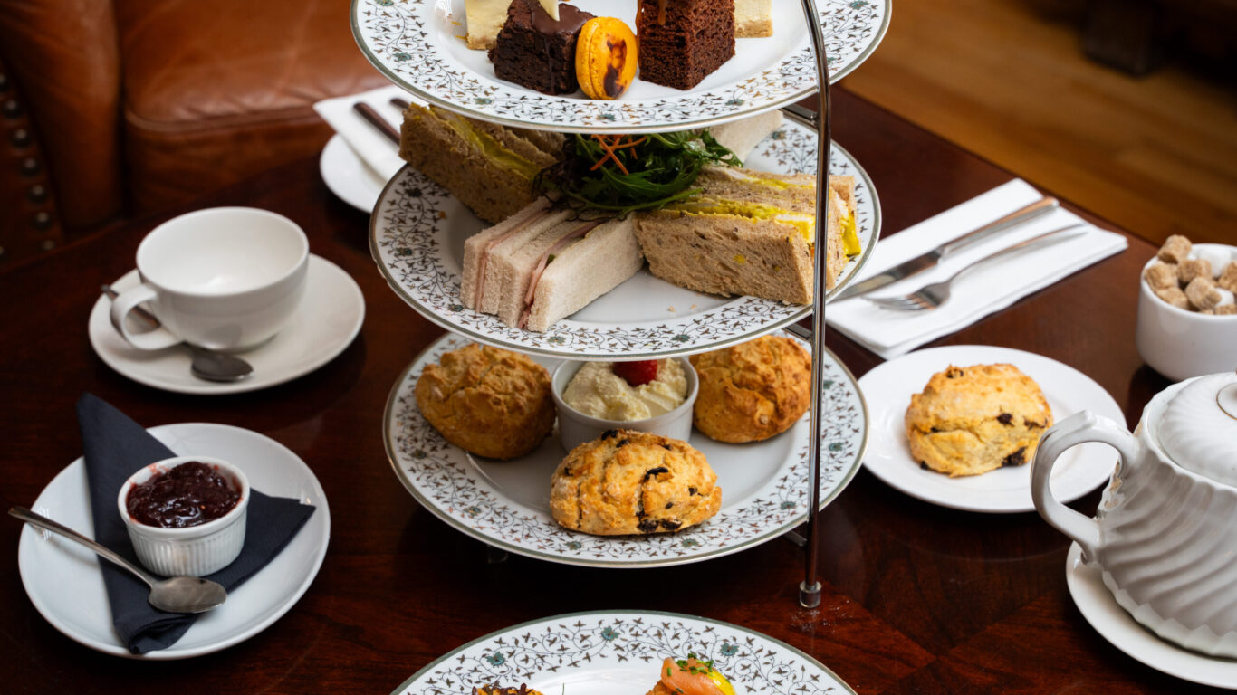 A tiered tray holds assorted finger sandwiches, scones, and pastries, set on a wooden table with teacups, a teapot, and a bowl of jam.