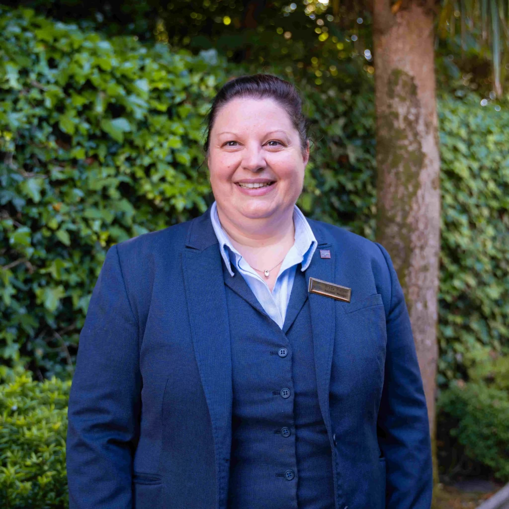 A woman in a dark blue suit stands outdoors in front of greenery, smiling at the camera.