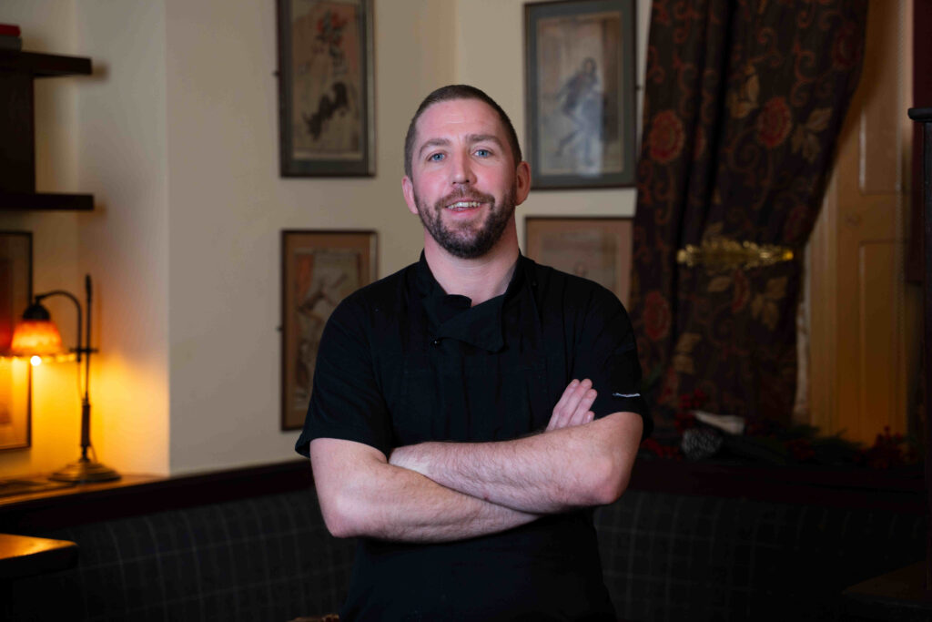 A man with short hair and a beard, wearing a black shirt, stands indoors with his arms crossed, in front of framed pictures on the wall.