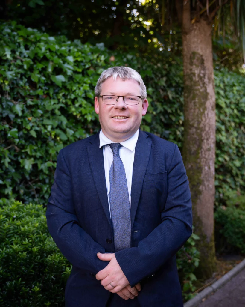 A man in a navy suit and tie stands outdoors in front of a leafy green hedge and a tree, smiling at the camera.