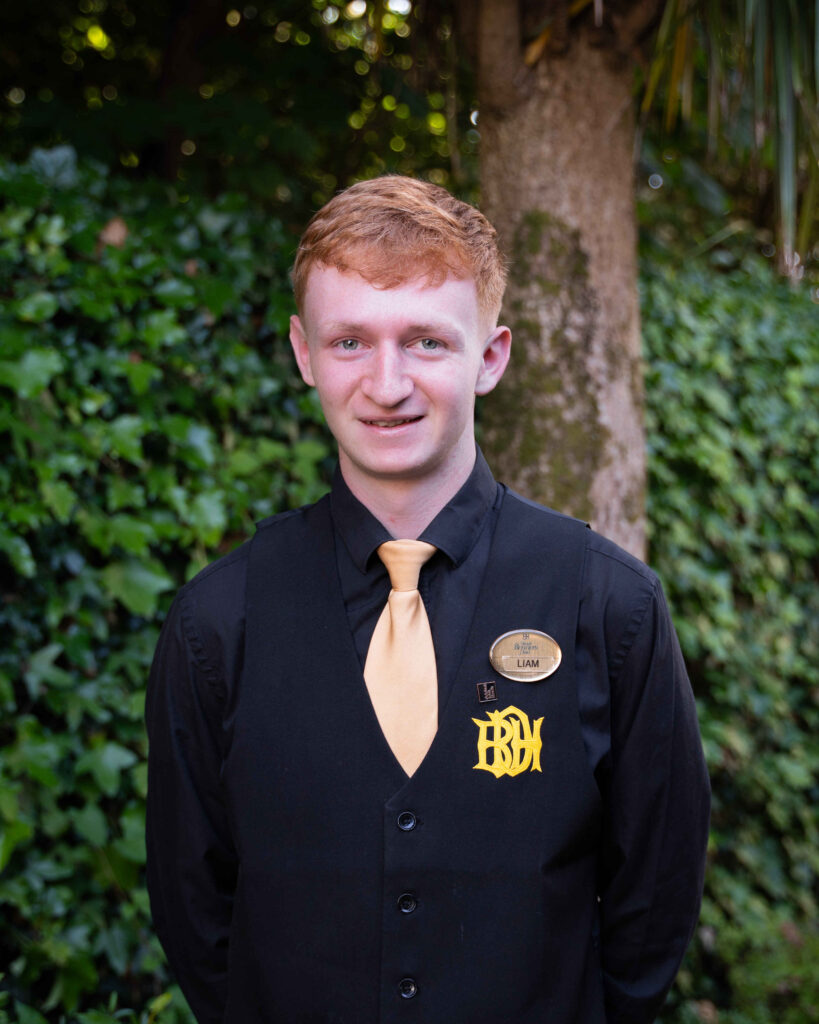 A young man with red hair wearing a black shirt, black vest with a yellow tie, and a name tag stands outdoors in front of greenery.