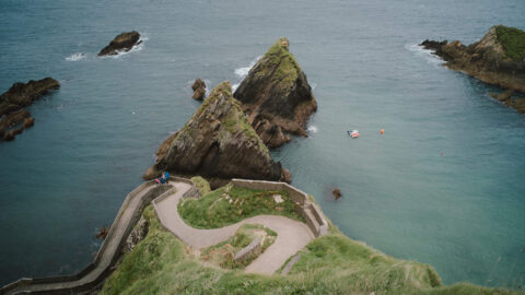 A winding stone pathway leads down a grassy cliff to the sea, with jagged rocks and a small boat floating in the water below.