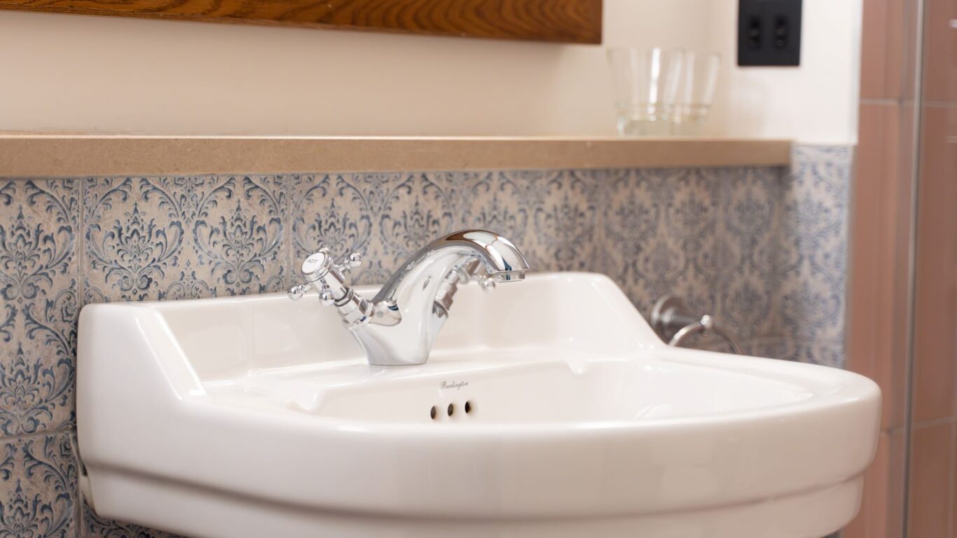 White ceramic sink with chrome faucet, patterned blue and white tile backsplash, wall-mounted mirror, and two clear glasses placed on the counter.