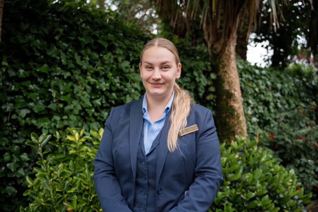 A woman in a blue suit with a name tag stands outdoors in front of green foliage and trees, smiling at the camera.