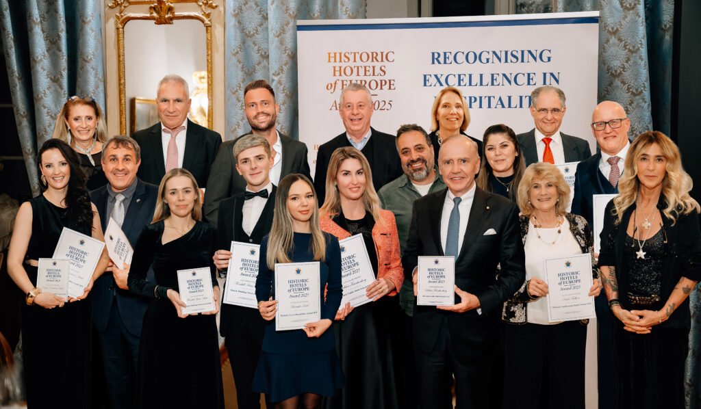 Gruppe A group of people in formal attire pose indoors, holding certificates in front of banners that read “Recognising Excellence in Hospitality” and “Historic Hotels of Europe Awards 2023.”.