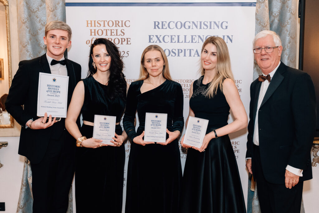 IMF_0480 Five people dressed formally stand in front of a banner holding award plaques at a Historic Hotels of Europe event.