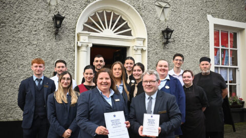 A group of hotel staff stand in front of Benner's Hotel entrance, with two people in front holding certificates and smiling at the camera.