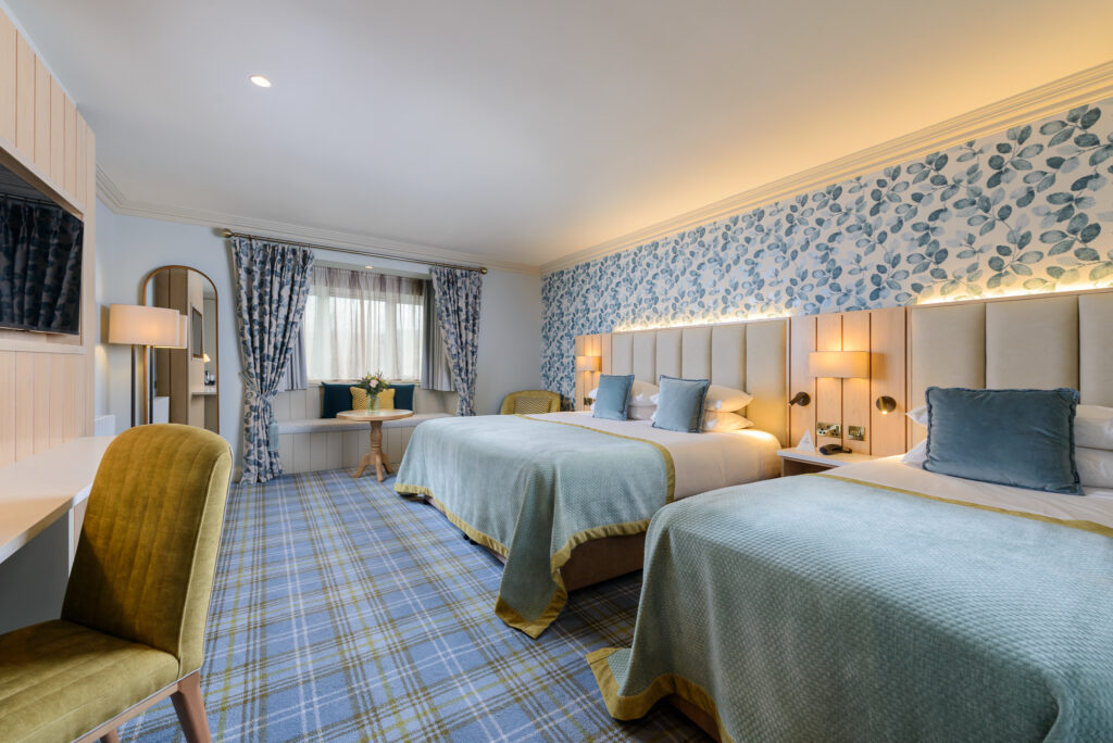 Hotel room with two double beds, blue and green decor, patterned wallpaper, a desk, a window with curtains, and a small table with flowers.