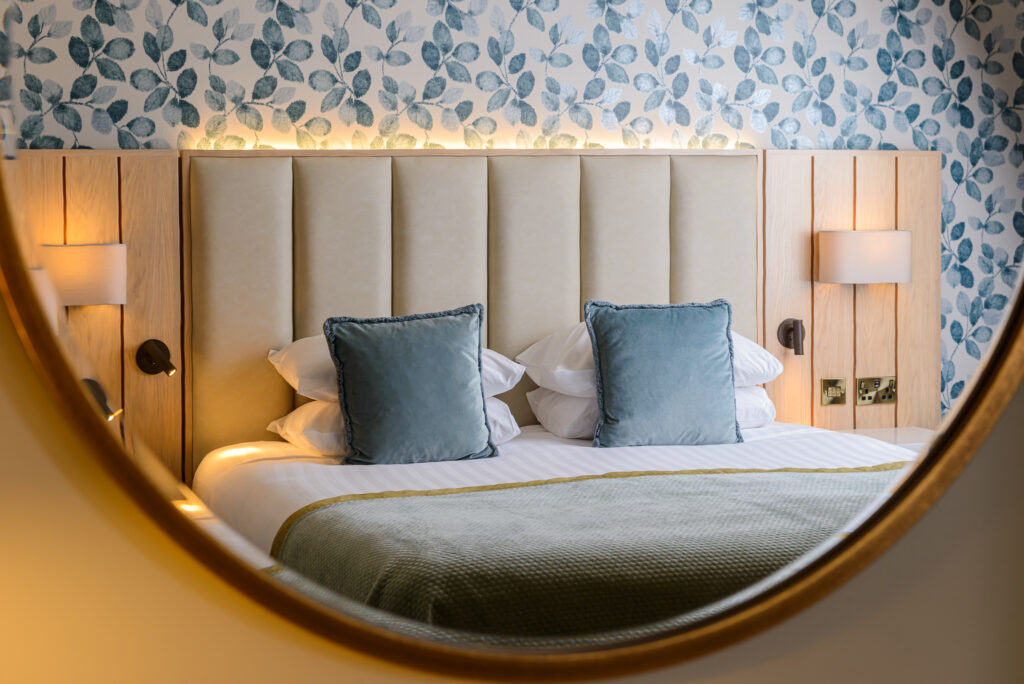 A neatly made bed with blue cushions and a green throw, photographed through a round mirror, with floral wallpaper and wall-mounted lights in the background.