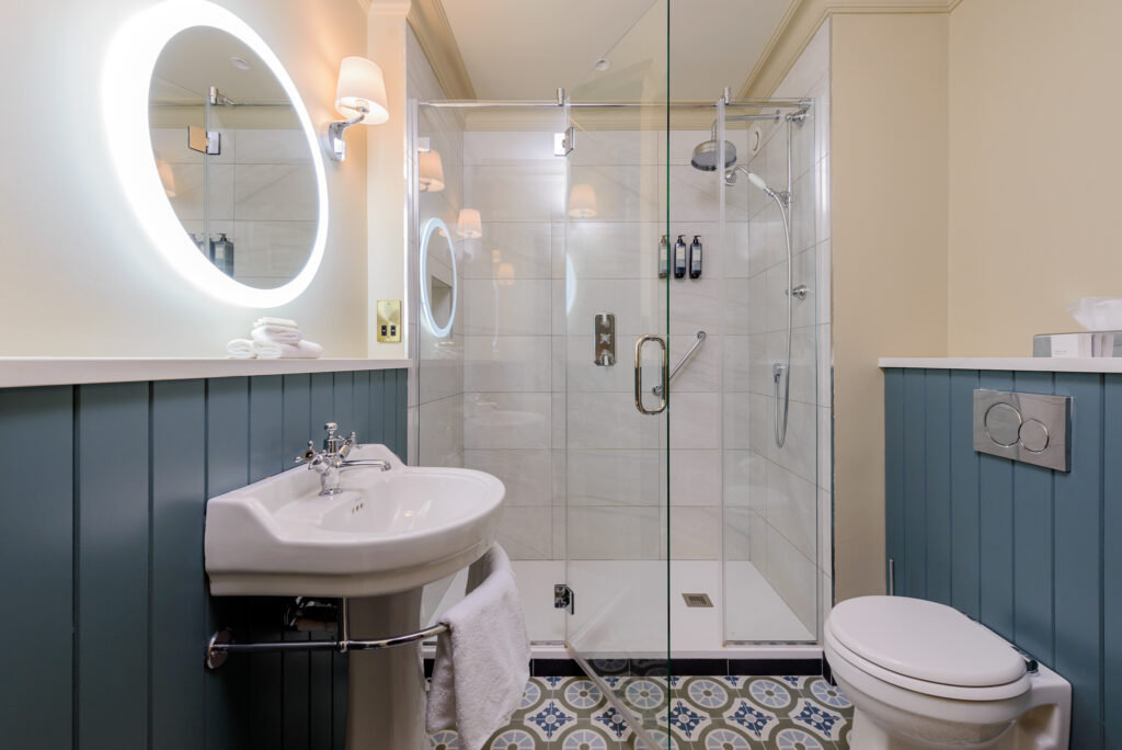 Modern bathroom with a glass-enclosed shower, illuminated round mirror above a sink, toilet, blue wainscoting, and patterned tile floor.