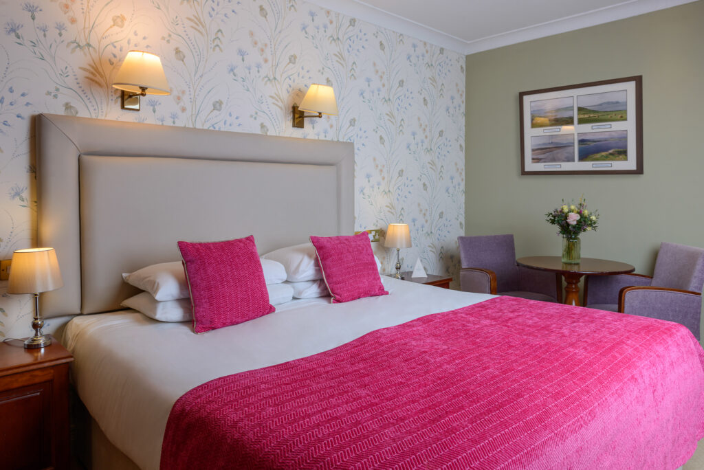 A hotel room with a double bed, pink pillows and blanket, floral wallpaper, two armchairs, a small round table with flowers, and framed art on the wall.