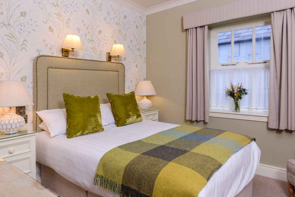 A neatly made double bed with green pillows and a checkered blanket, two bedside tables with lamps, floral wallpaper, and a window with curtains and a vase of flowers.