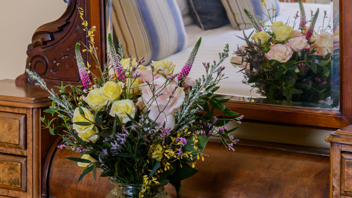 A glass vase with a mixed flower bouquet sits on a wooden vanity with a large mirror reflecting a bed and pillows in the background.