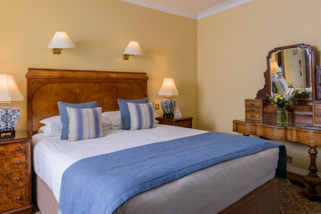 A neatly made double bed with blue and white bedding, two bedside tables with lamps, and a wooden vanity with a mirror and flowers in a cozy, well-lit bedroom.