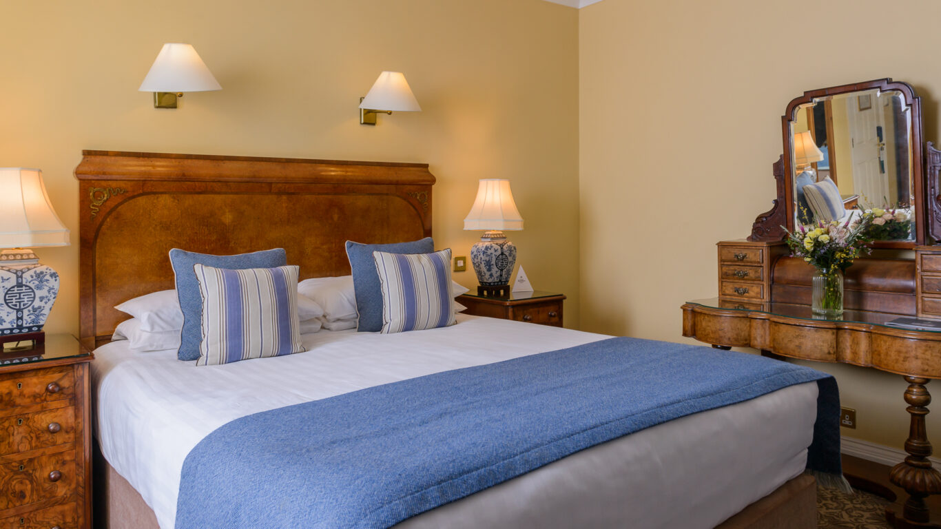 A neatly made double bed with blue and white bedding, two bedside tables with lamps, and a wooden vanity with a mirror and flowers in a cozy, well-lit bedroom.