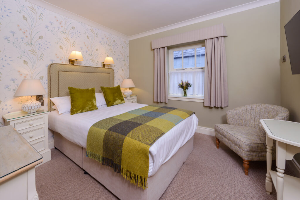 A neatly made double bed with green accents, flanked by nightstands and lamps, sits in a cozy, carpeted bedroom with a window, armchair, and TV.