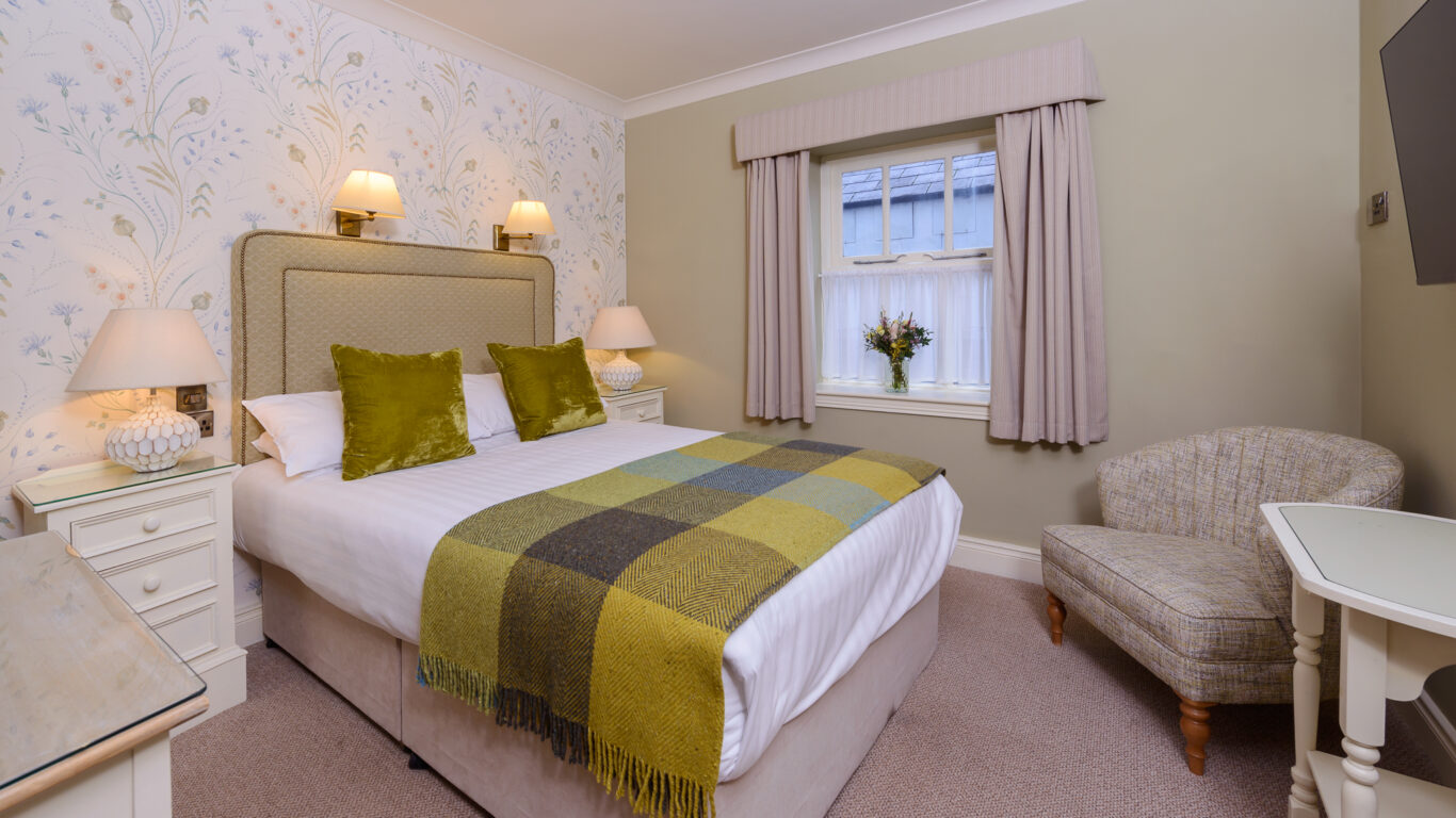 A neatly made double bed with green accents, flanked by nightstands and lamps, sits in a cozy, carpeted bedroom with a window, armchair, and TV.