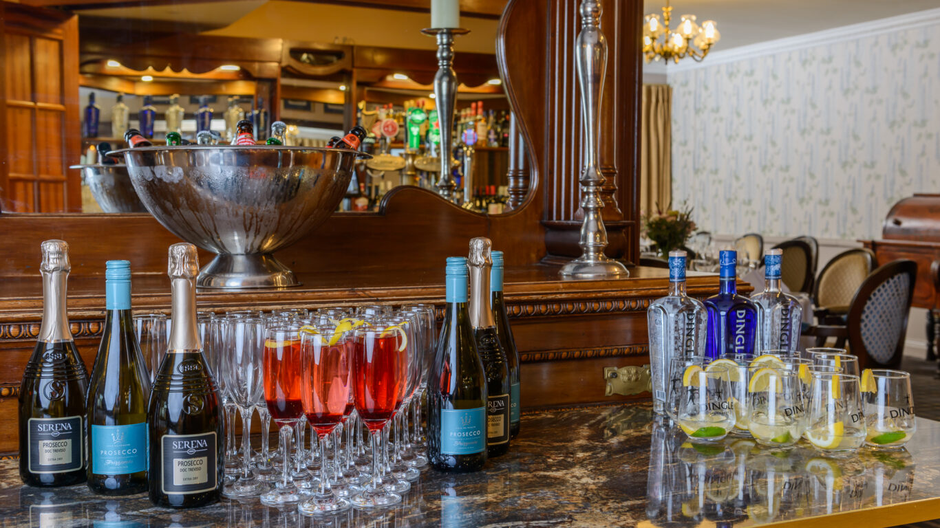 Bottles of sparkling wine, glasses with red drinks, bottles of gin, and gin and tonics with lemon slices are arranged on a marble bar counter in an elegant setting.