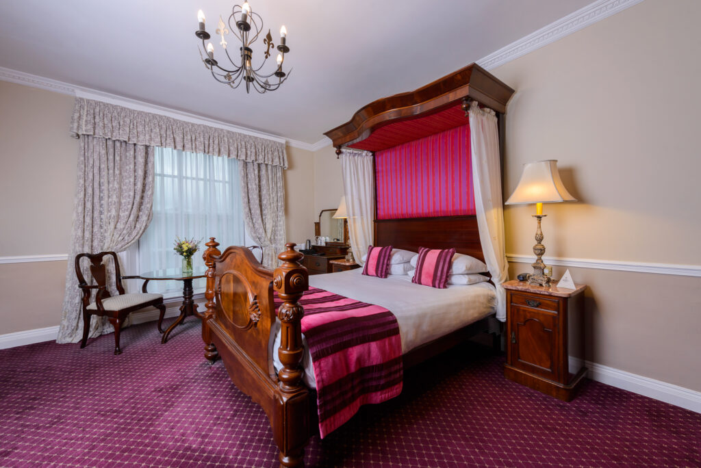 A traditional bedroom with a wooden canopy bed, pink and burgundy bedding, a chandelier, a side table with a lamp, and a window with drapes.