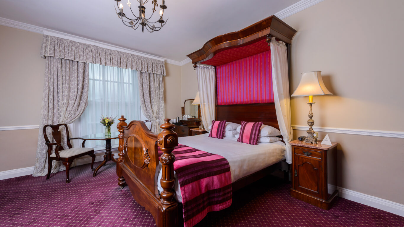 A traditional bedroom with a wooden canopy bed, pink and burgundy bedding, a chandelier, a side table with a lamp, and a window with drapes.