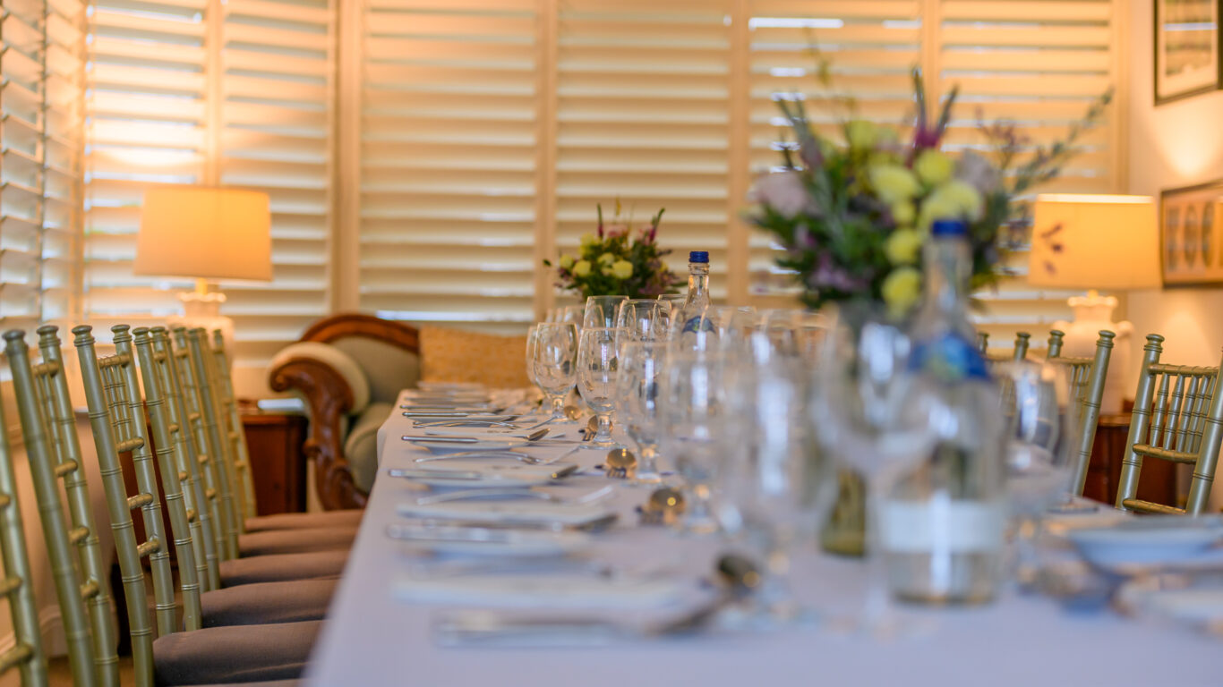 A long dining table set with glassware, plates, cutlery, and floral centerpieces in a well-lit room with shutters and gold chairs.