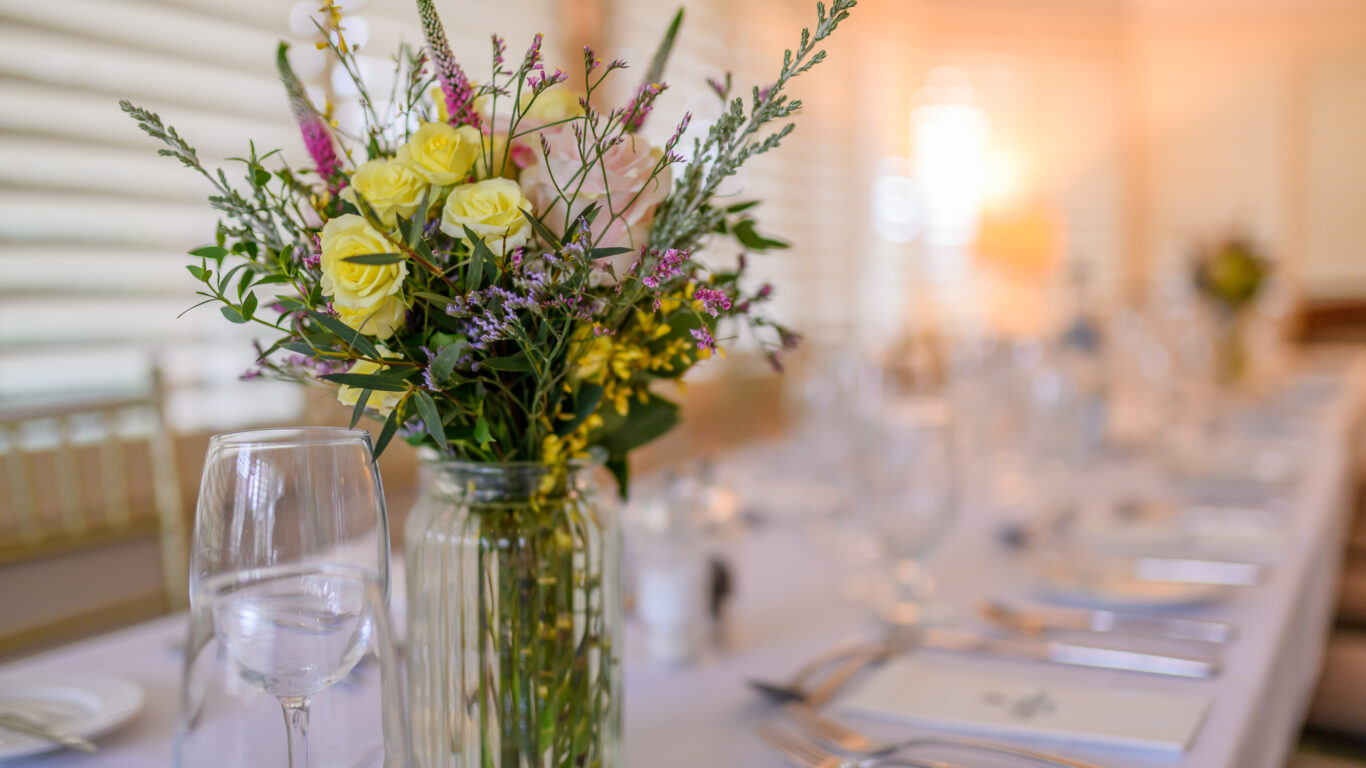 A glass vase with yellow and purple flowers sits on a set dining table with wine glasses, cutlery, and plates in a well-lit room.
