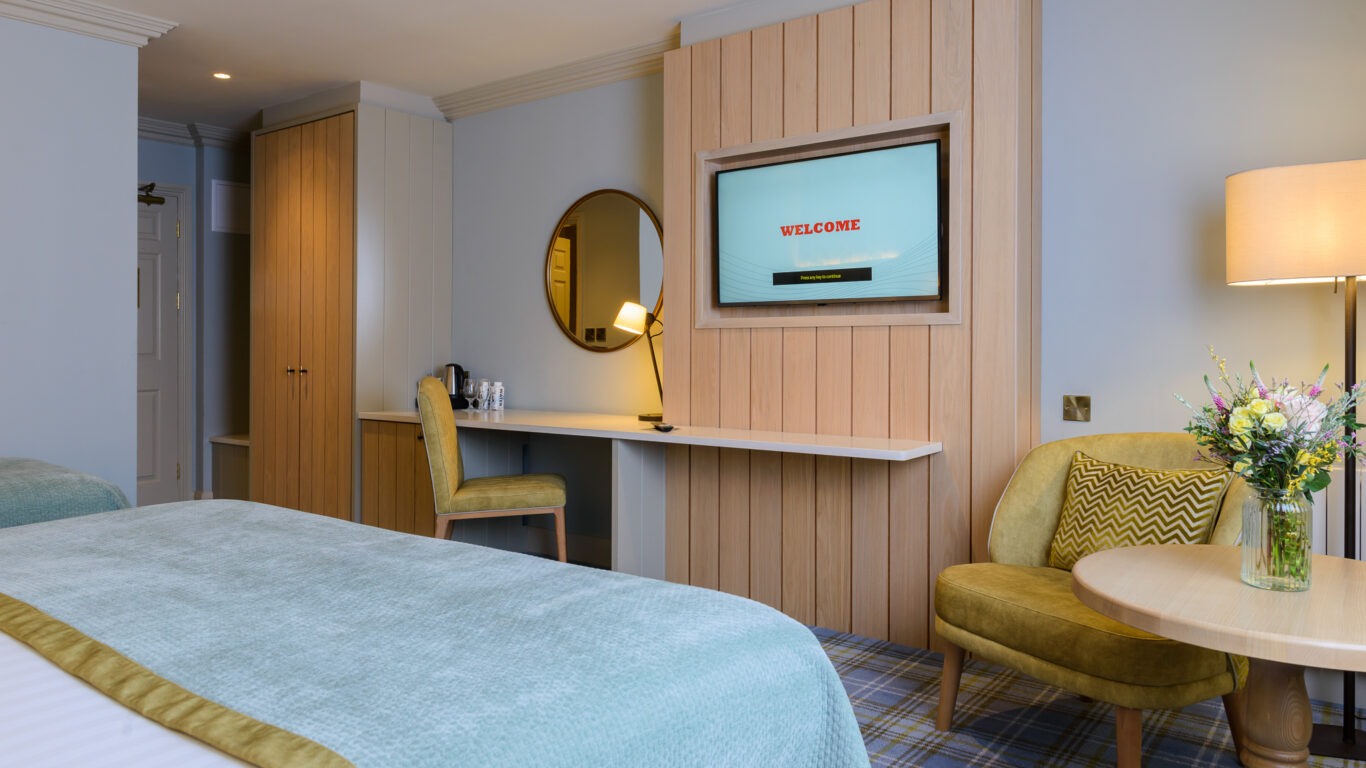 A modern hotel room with a bed, a desk and chair, a wall-mounted TV displaying 