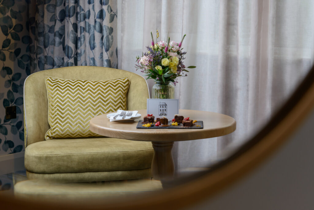A round wooden table with a vase of flowers, a plate of desserts, and a napkin, next to a cushioned chair with a chevron-patterned pillow.