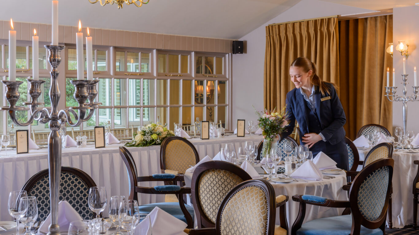 A waitress in formal attire arranges flowers on a table in an elegantly set dining room with candelabras, white linens, and neatly arranged glassware.