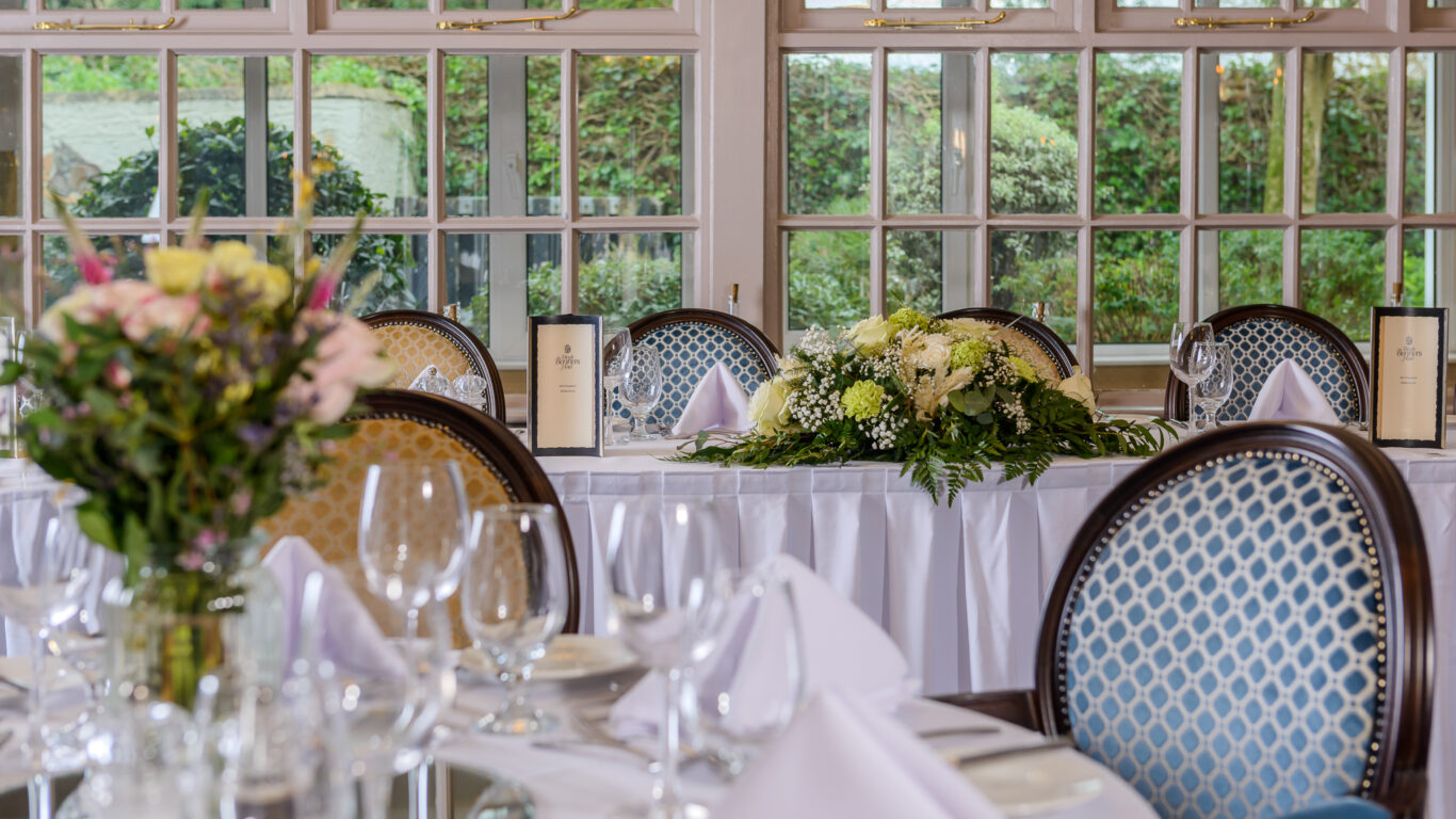 A formal dining table is set with white linens, glassware, floral centerpieces, menus, and decorative chairs in a room with large windows overlooking a garden.