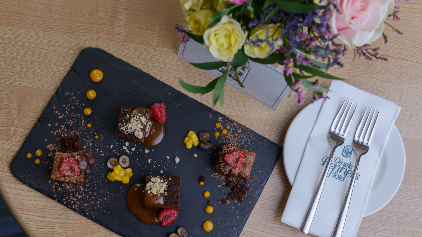 A slate platter with three chocolate desserts, garnished with raspberries, sauce, and crumbs, beside a plate, napkin, cutlery, and a bouquet of flowers on a wooden table.