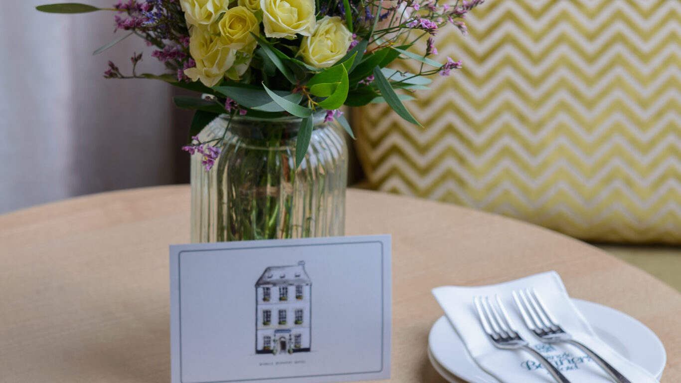 A slate platter with assorted chocolate desserts and fruit puree is on a round table, next to a vase of flowers, a card, and a plate with forks and napkins.