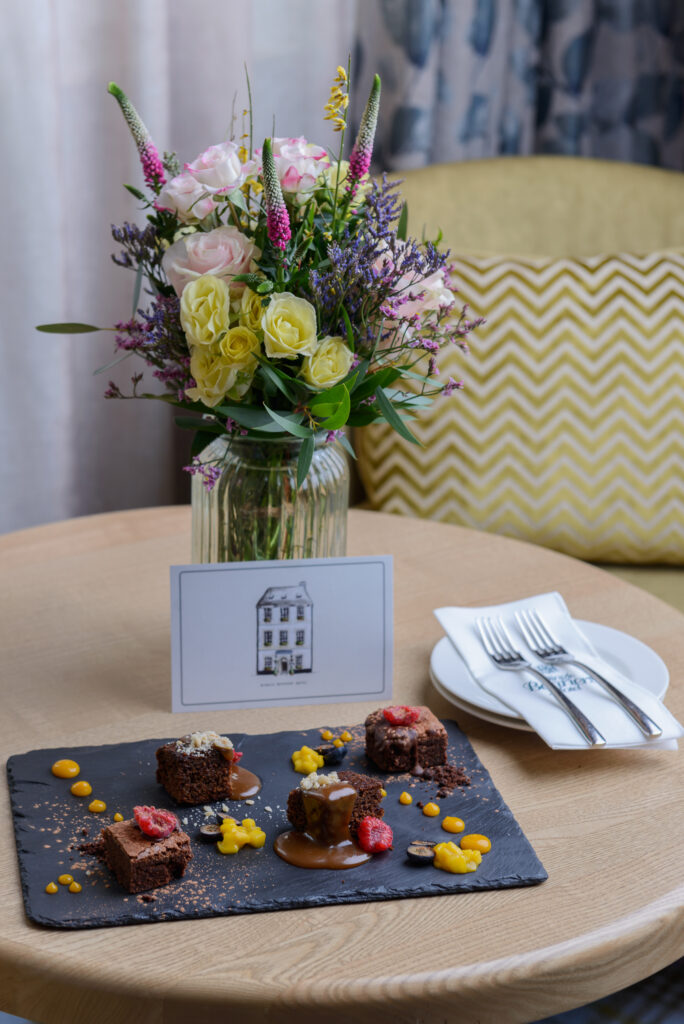 A slate platter with assorted chocolate desserts and fruit puree is on a round table, next to a vase of flowers, a card, and a plate with forks and napkins.
