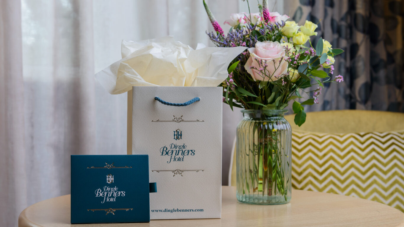 A Dingle Benners Hotel gift bag and envelope sit on a round wooden table next to a glass vase of assorted flowers, with a zigzag-patterned chair in the background.
