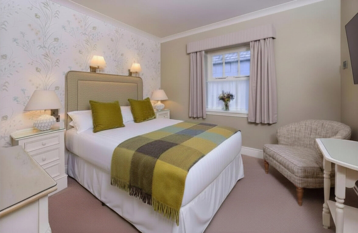 A neatly made double bed with green pillows and a plaid blanket in a cozy, softly lit bedroom with a chair, two nightstands, and a window with beige curtains.